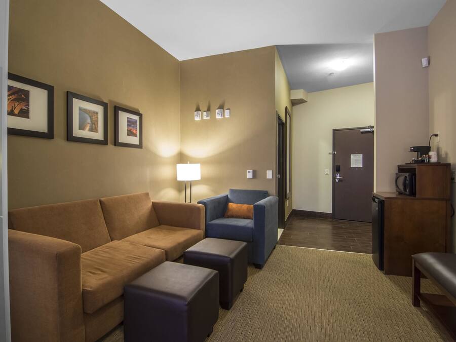 Habitación de hotel con sofá marrón, sillón azul, una mesita pequeña y un banco. Hay una lámpara, un refrigerador y un microondas sobre un armario de madera.