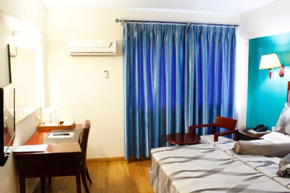 A hotel room with a bed, a desk, a chair, a small table, and a window with blue curtains.