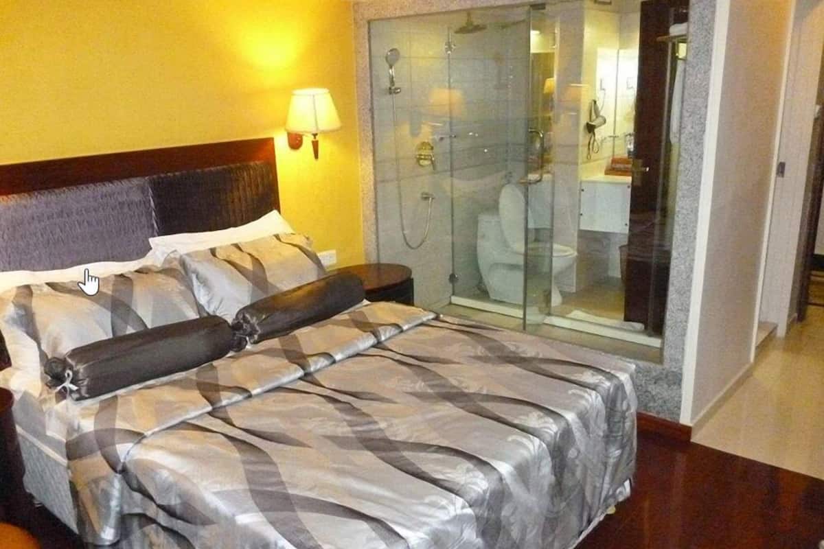 A hotel room with a bed, a nightstand, a lamp, a glass-enclosed shower, and a bathroom visible through the glass.
