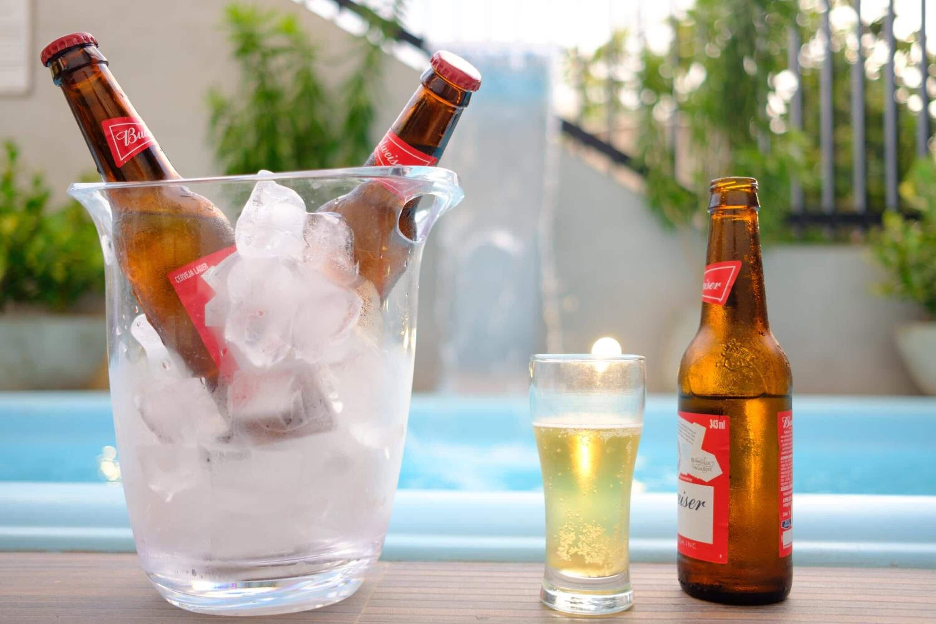 Una cerveza en un vaso junto a una botella y un vaso medio lleno sobre una mesa de madera junto a una piscina.