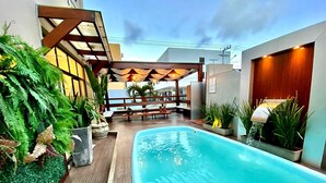 Une piscine dotée d’une terrasse en bois, entourée de plantes en pot et agrémentée d’un espace salon abrité.