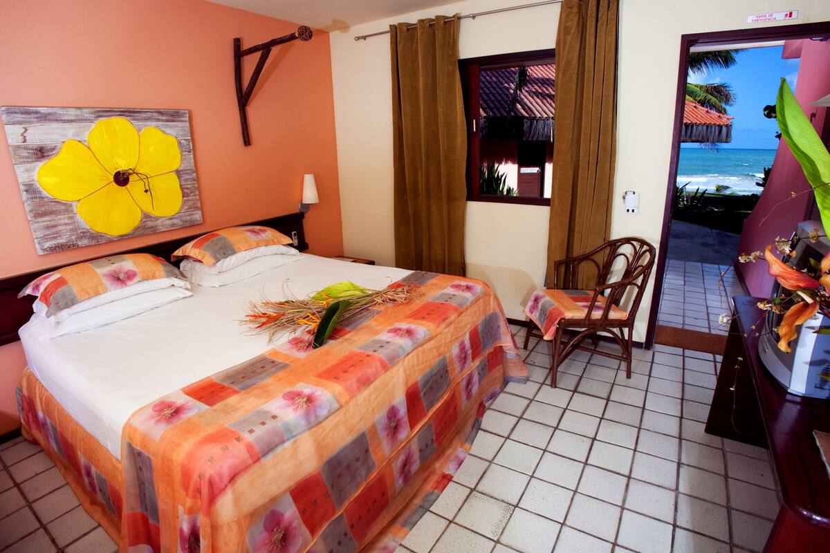 Habitación de hotel con cama, una silla, una mesa, un cuadro de una flor y vista al océano a través de una ventana.