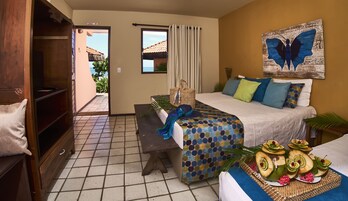 A hotel room with a bed, a desk, a bench, a dining table with fruit and flowers, a large window, and a view of the outside.