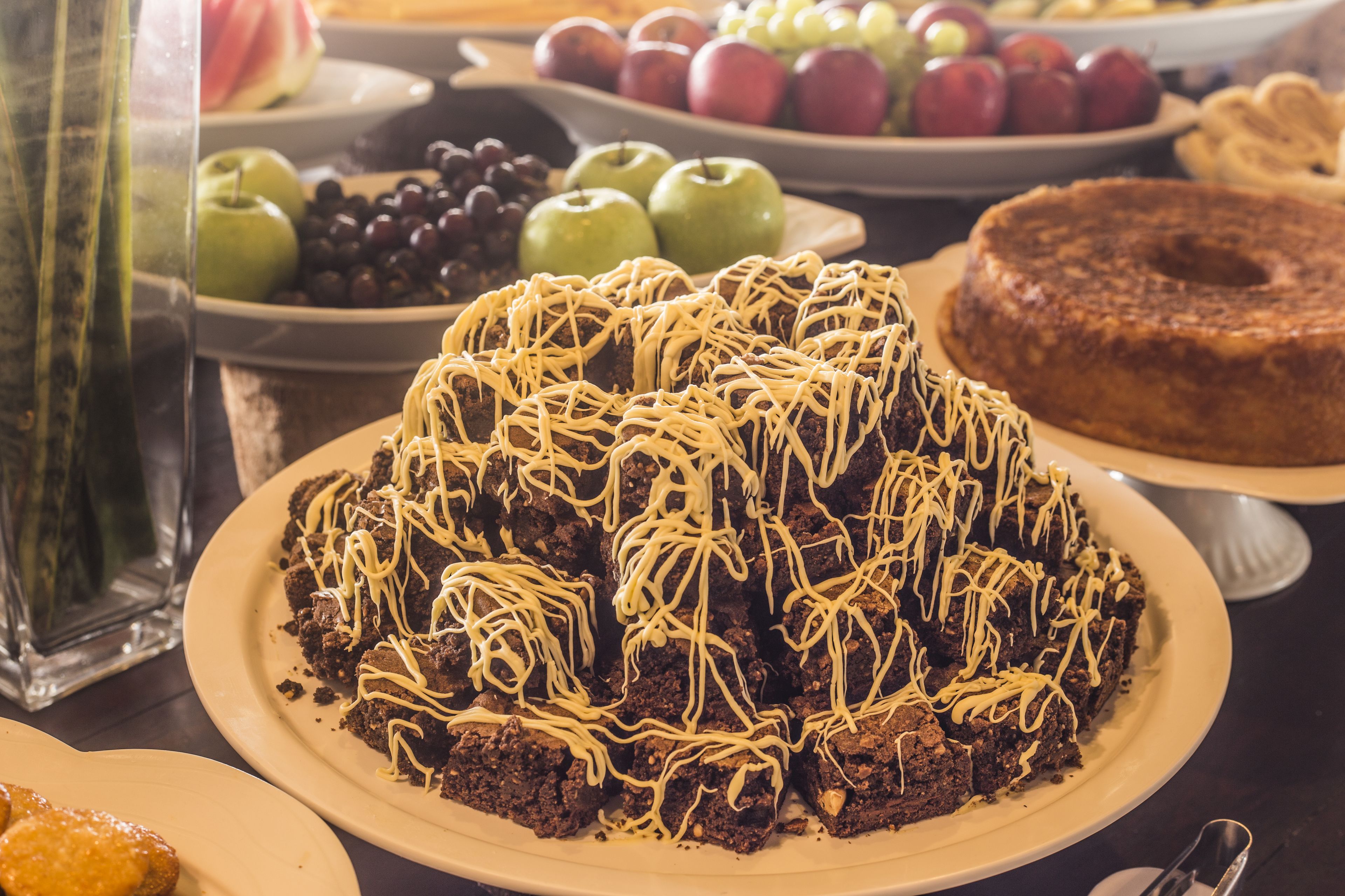 A dessert table with various sweets including a chocolate cake with white icing, plates of apples, grapes, and a bundt cake.