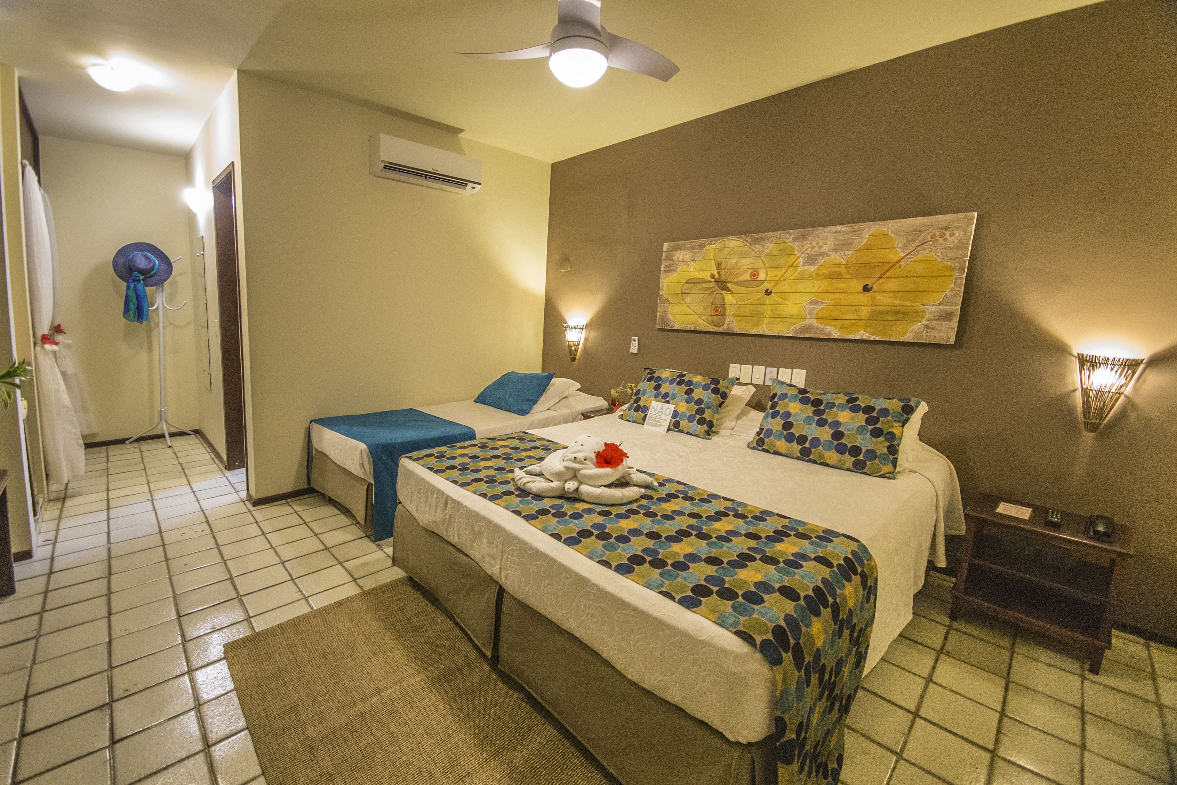 Een hotelkamer met een groot bed, een klein bureau, een stoel, een ventilator, een airconditioner aan de muur en een schilderij.