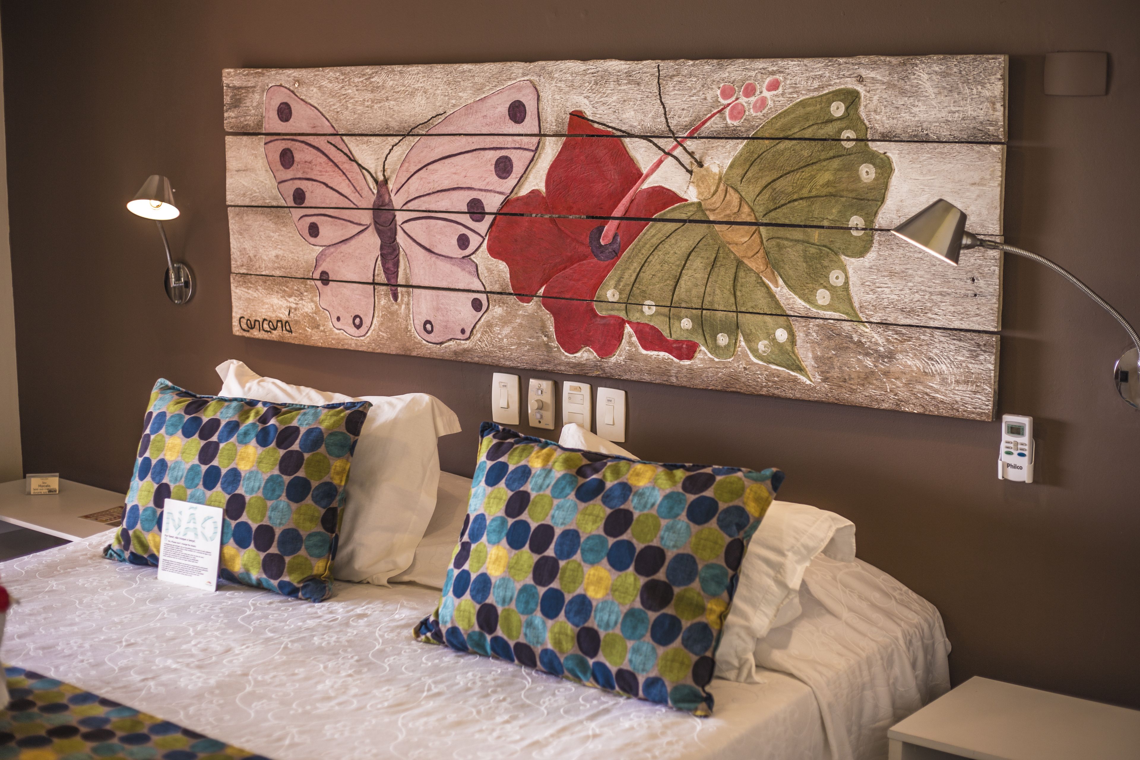 A hotel room with a bed, two pillows, a wall-mounted lamp, and a large butterfly and flower painting.