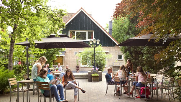 An outdoor patio area with tables and chairs, surrounded by greenery and a building in the background. - Stayokay Arnhem - Hostel (Arnhem)