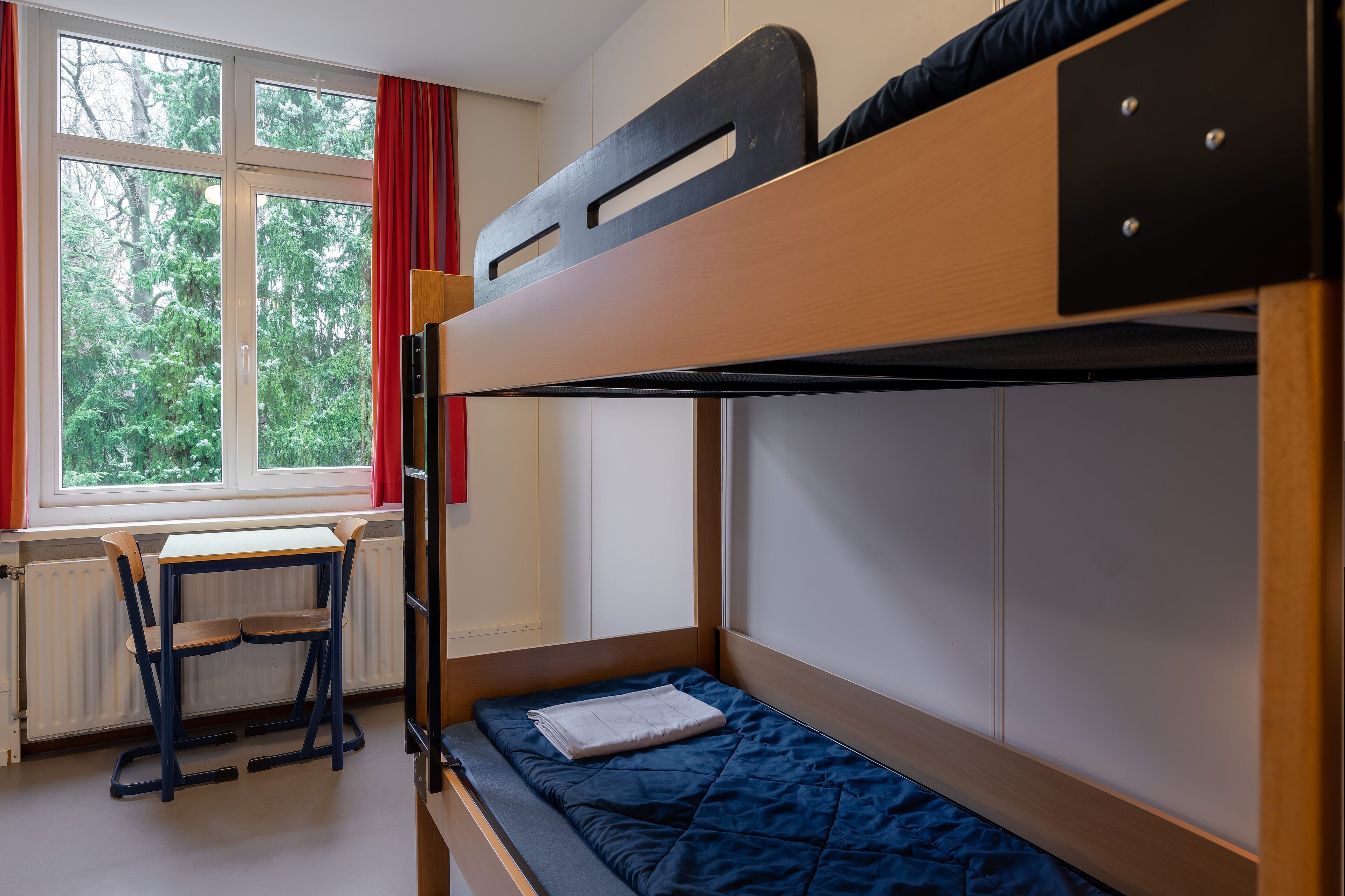 A bunk bed with a desk and chair, a window with red curtains, and a view of greenery outside.