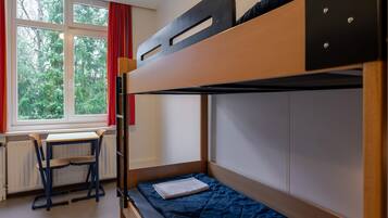A bunk bed with a desk and chair, a window with red curtains, and a view of greenery outside.