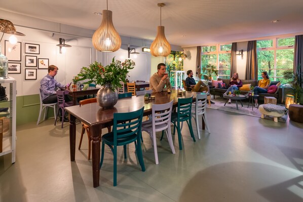 A modern dining area with wooden tables and chairs, large windows with curtains, and hanging pendant lights.