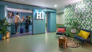 A hotel reception area with a blue wall, a reception desk, and a seating area with a couch and chairs. - Stayokay Arnhem - Hostel (Arnhem)
