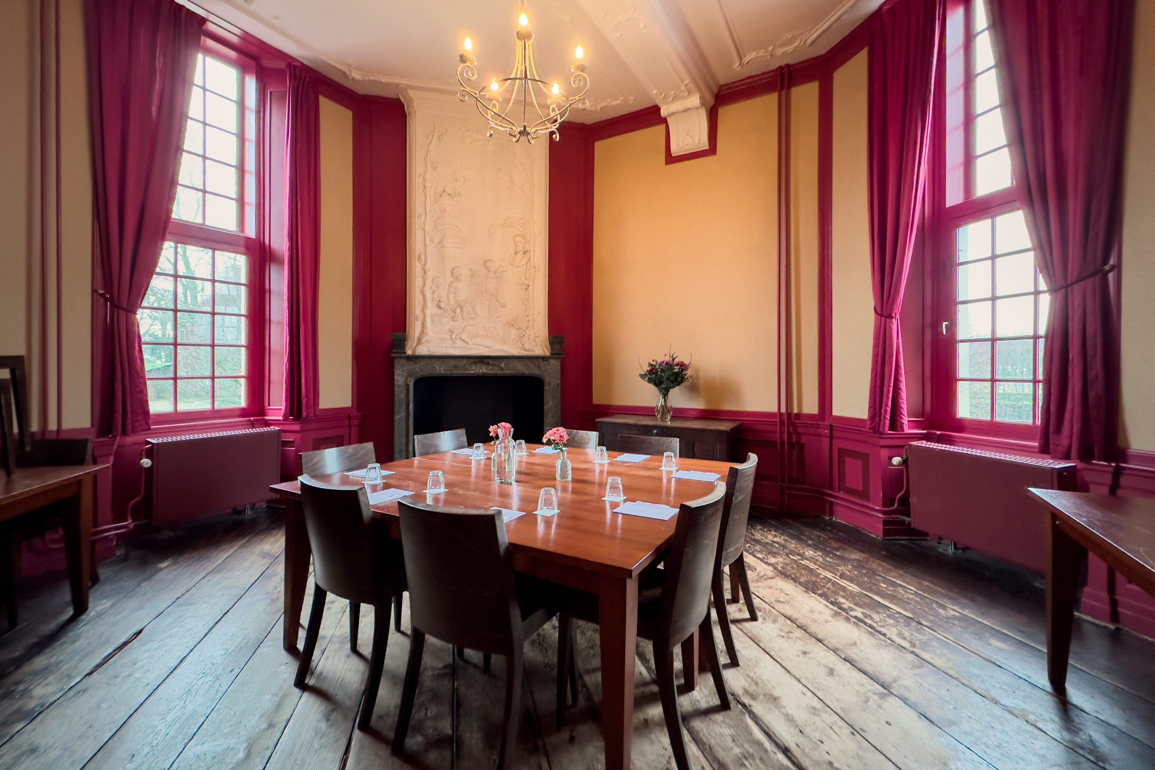 A dining room with a wooden table set for six, flanked by red curtains and a fireplace.