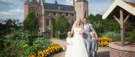 Un couple de jeunes mariés et deux enfants se tiennent devant un bâtiment historique doté d’un jardin.