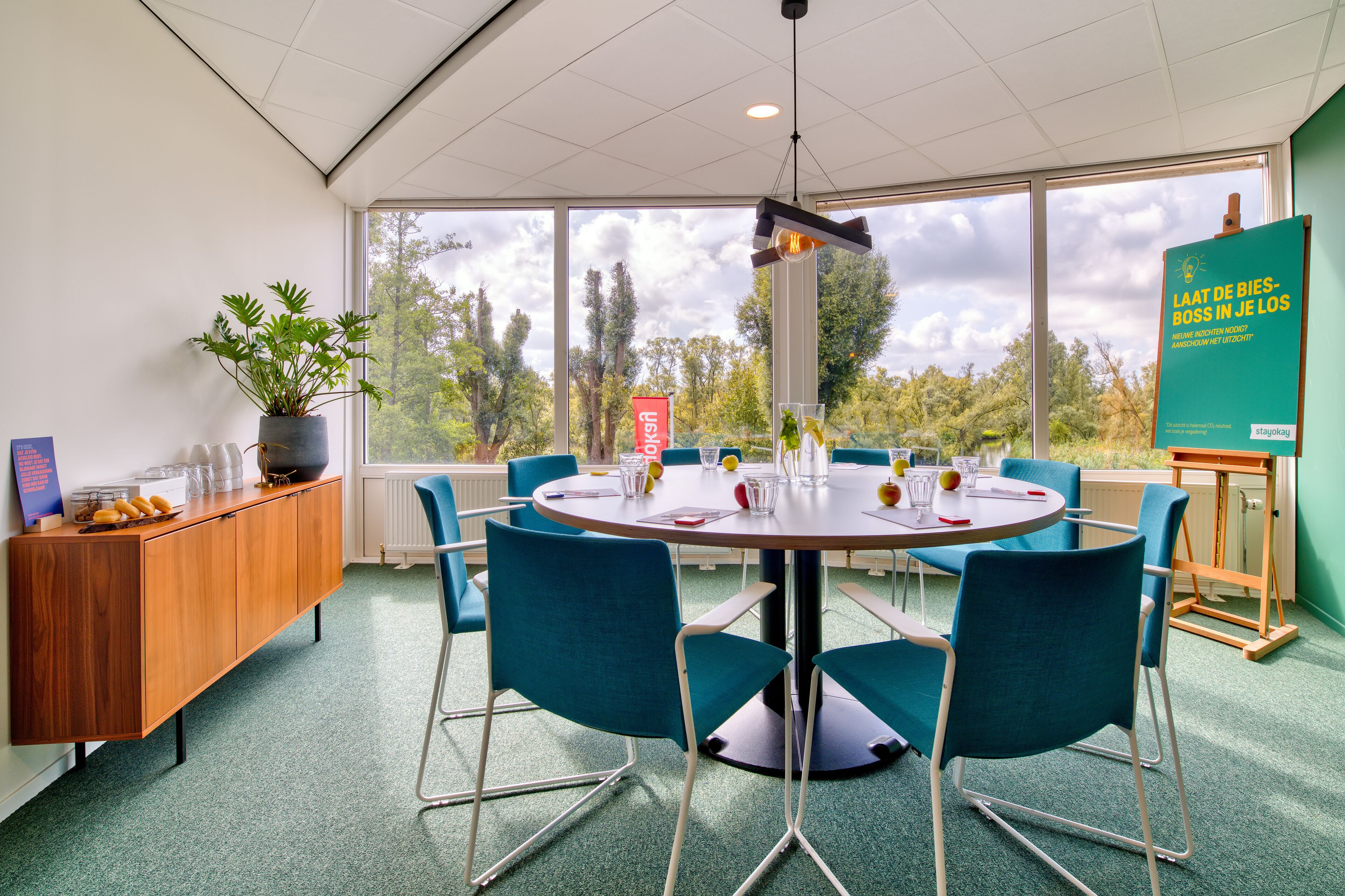 A conference room with a round table, blue chairs, a wooden cabinet, and a large window with a view of trees.