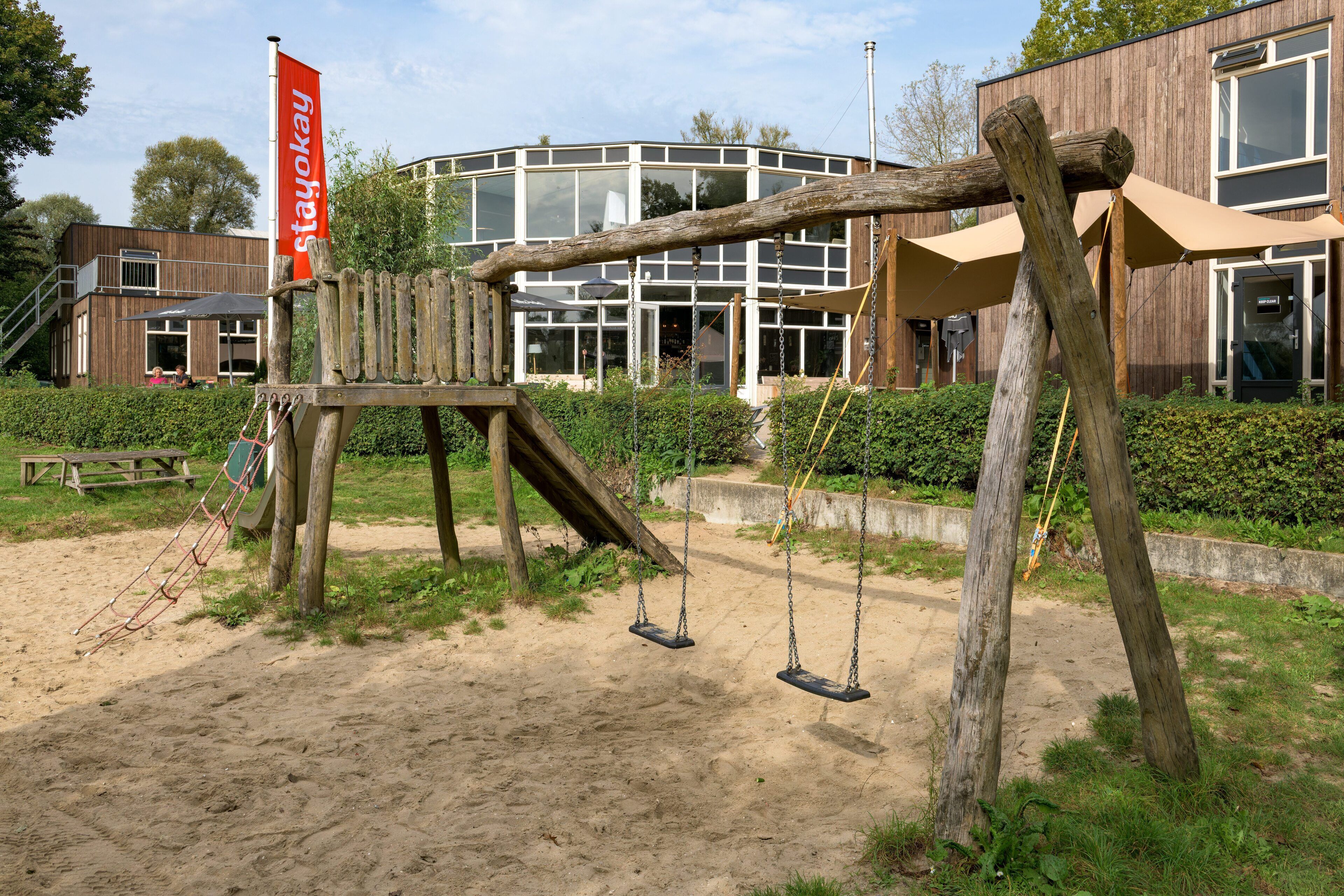 Een houten schommelset met een rode banner in een park, met een modern gebouw op de achtergrond.