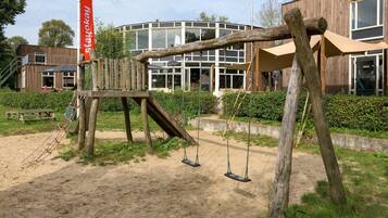Een houten schommelset met een rode banner in een park, met een modern gebouw op de achtergrond.