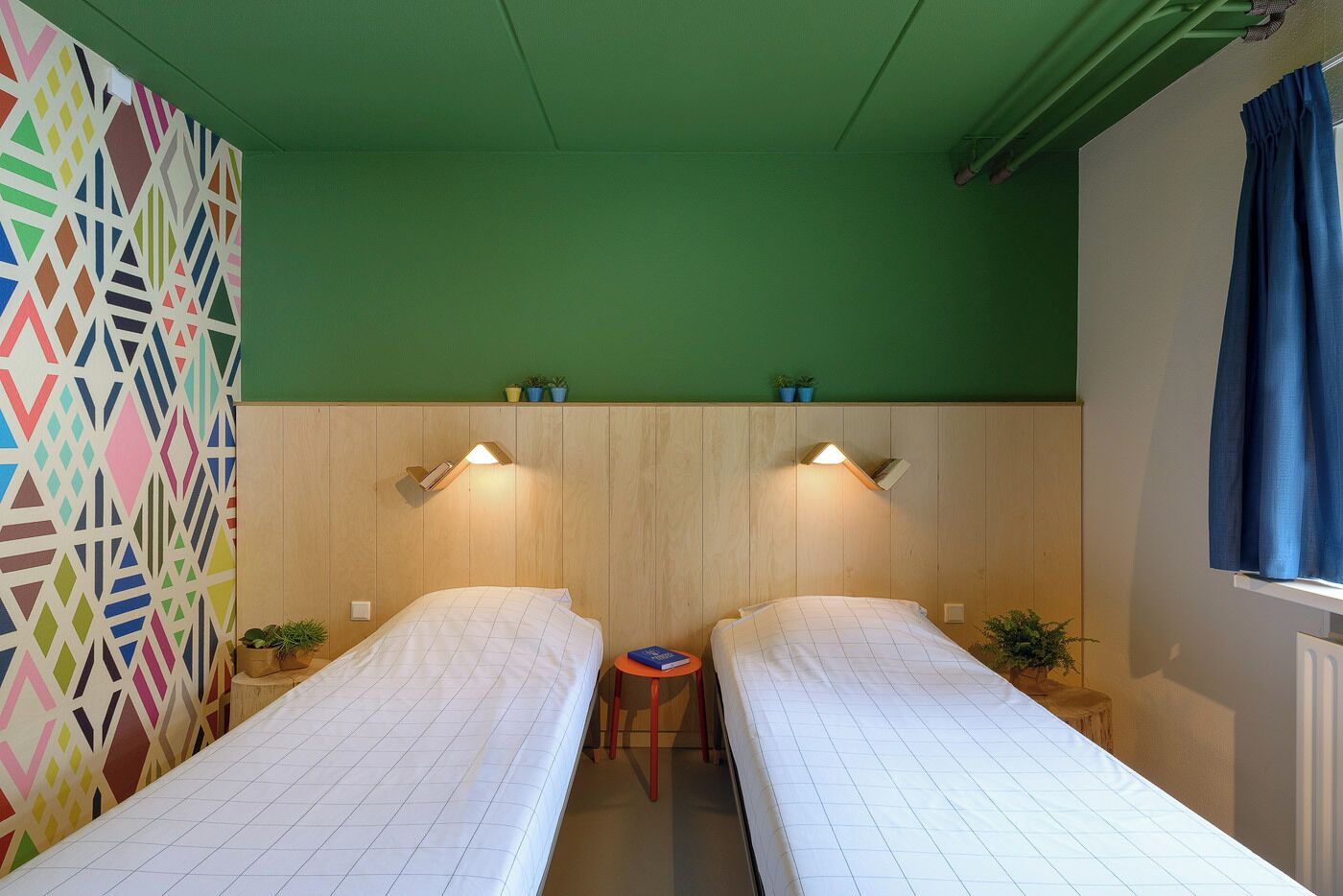 A room with two single beds, a small table, and a colorful geometric-patterned wall.