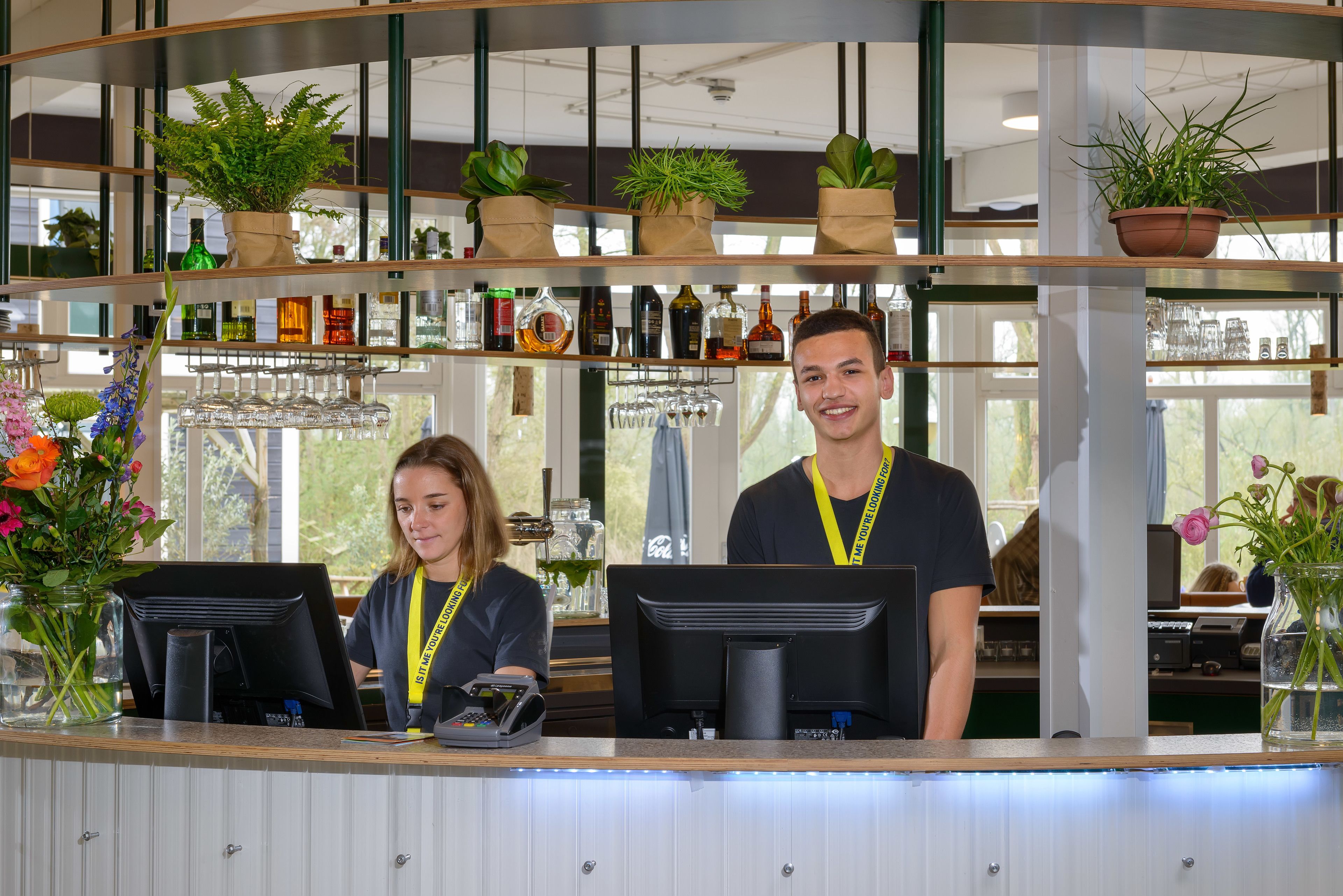 A bar area with two staff members, a counter with a payment terminal, shelves with bottles and glasses, and potted plants.