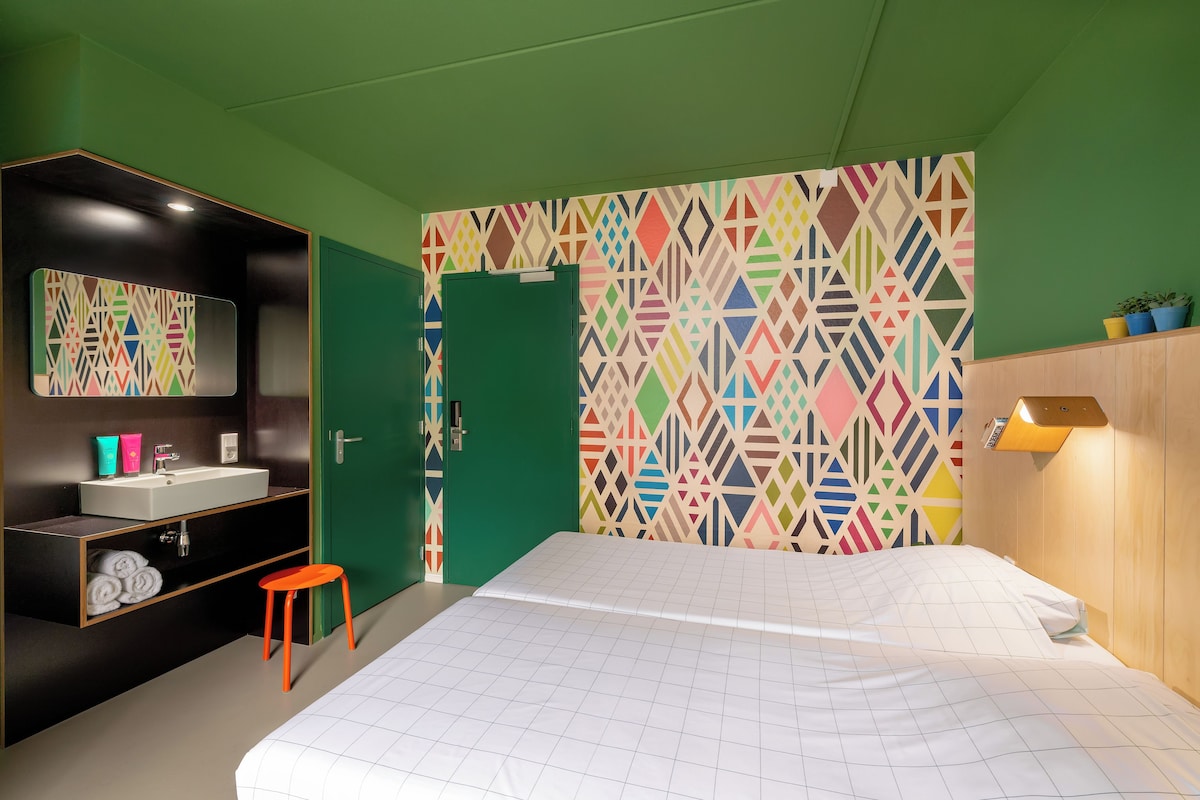 Een slaapkamer met een bed, een groene deur, een badkamer met een wastafel en een kleurrijk geometrisch patroon op de muur.