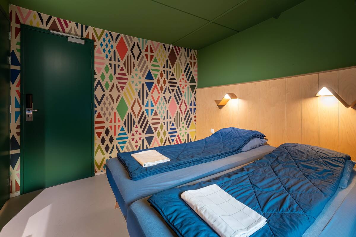 A room with two beds, a colorful geometric wall, a green door, and wooden paneling on the walls.