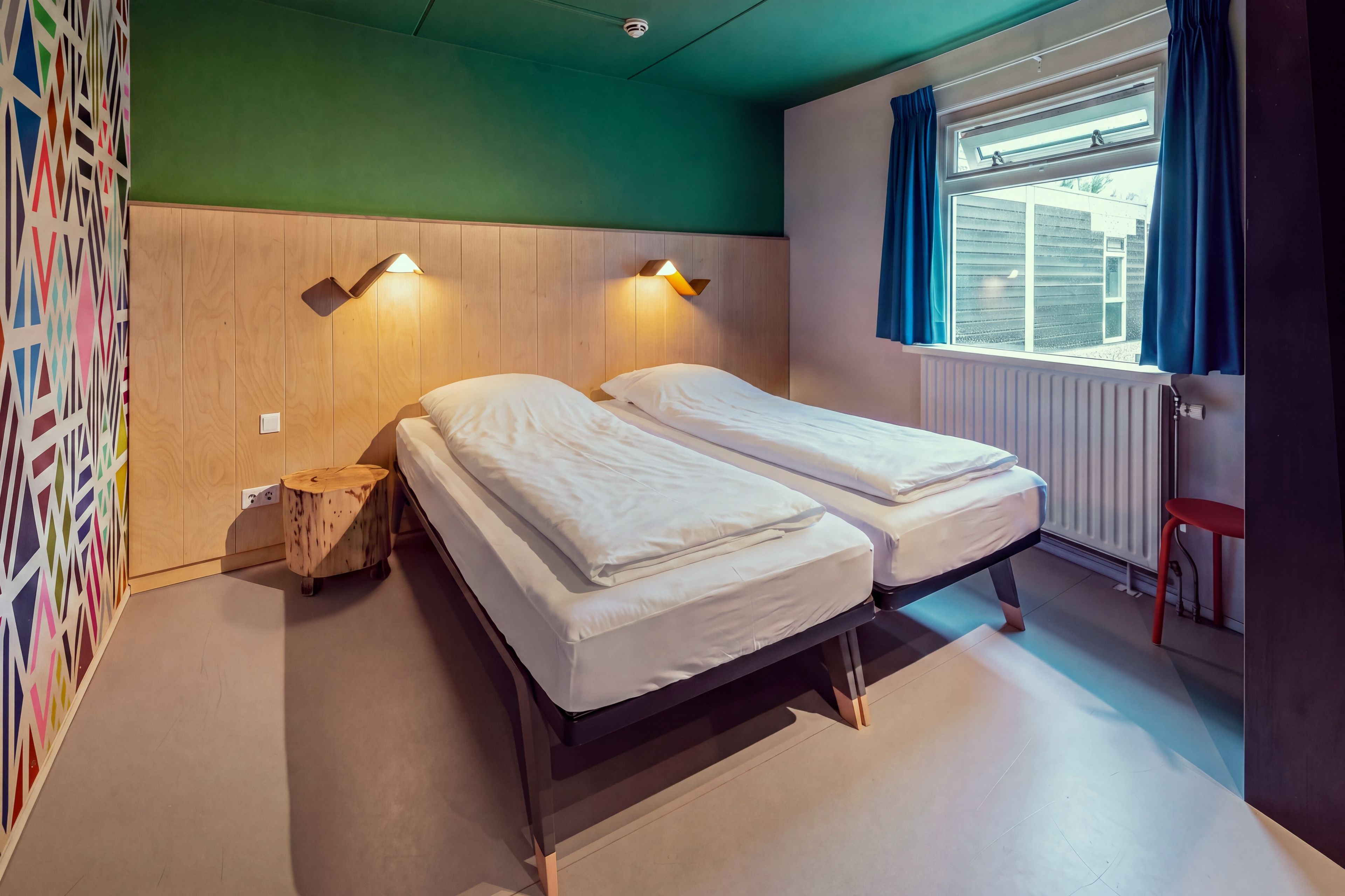 A room with two beds, a window with curtains, a radiator, and a wall with a geometric pattern.