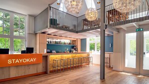 A modern hotel lobby with a reception desk labeled "STAYOKAY," a bar area, wooden flooring, and a mezzanine level with a railing and hanging light fixtures. - Stayokay Soest - Hostel (Soest)