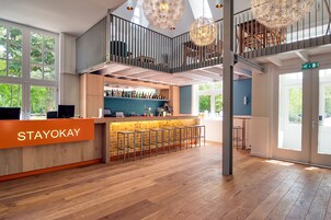 A modern hotel lobby with a reception desk labeled "STAYOKAY," a bar area, wooden flooring, and a mezzanine level with a railing and hanging light fixtures.