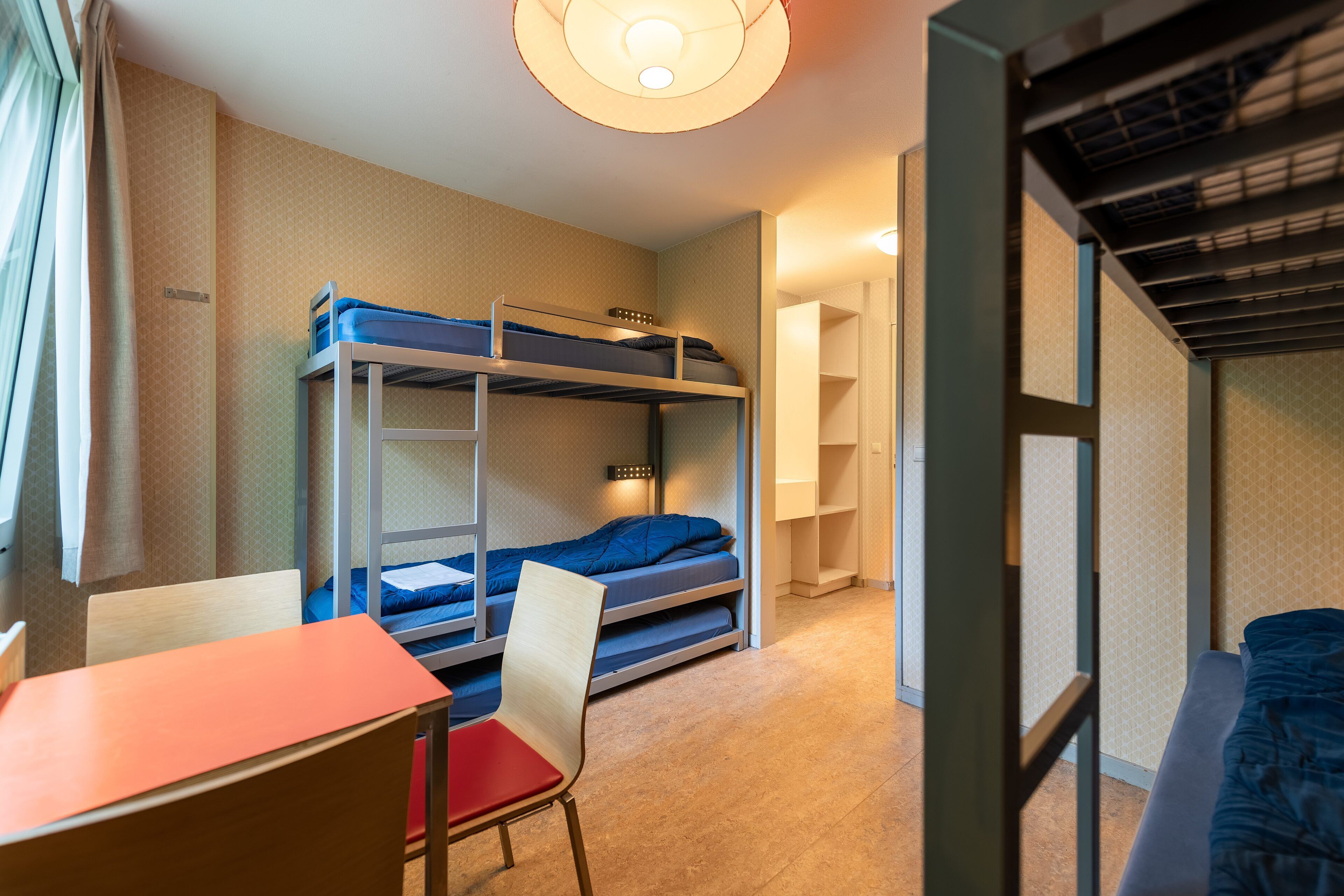 A bunk bed room with a desk and chair, a window with curtains, and a door leading to another room.