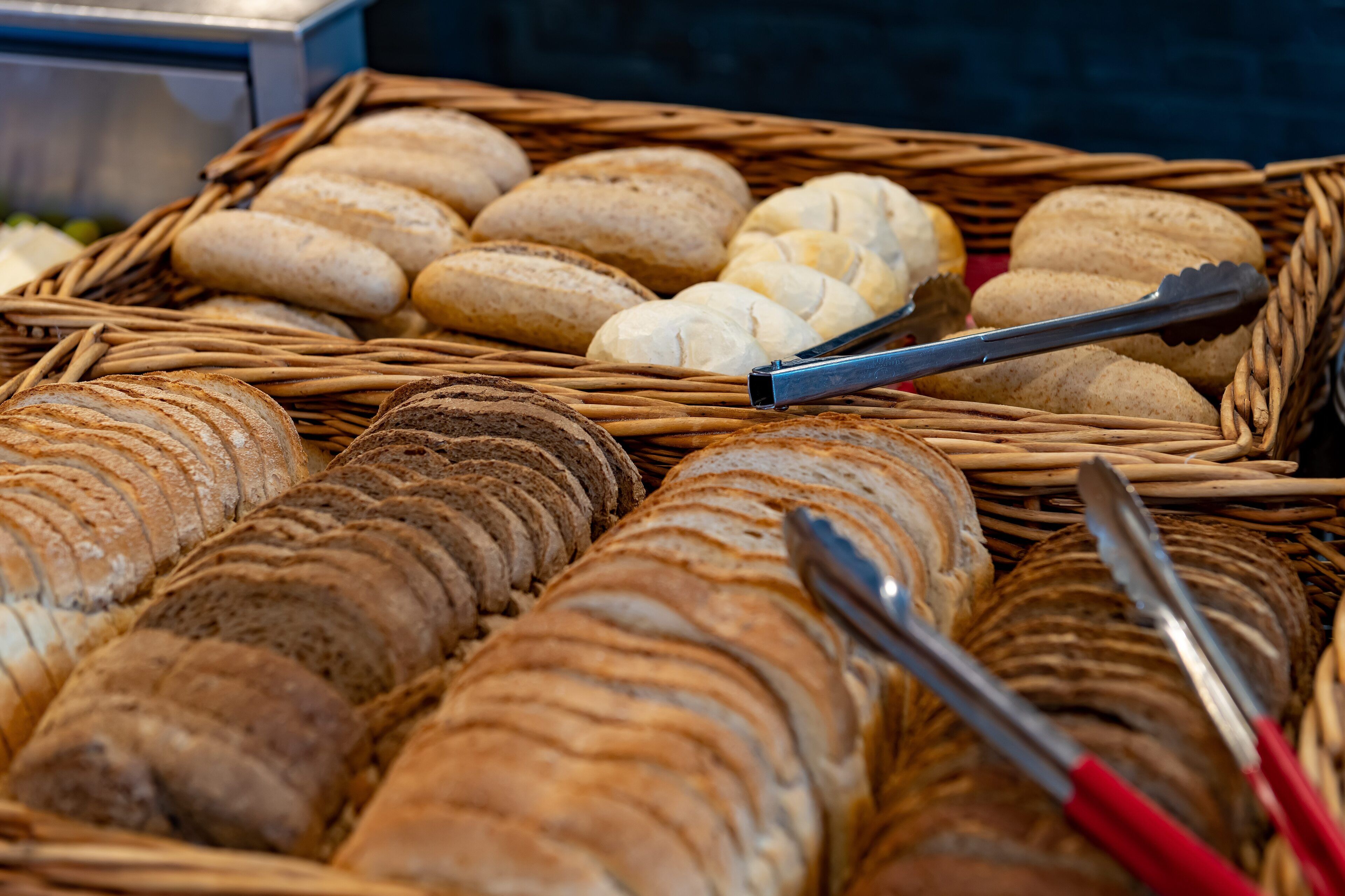 A variety of breads in a wicker basket with tongs.