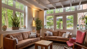 A cozy living room with two sofas, a coffee table, and a red armchair. Large windows offer views of the outdoors. - Stayokay Soest - Hostel (Soest)