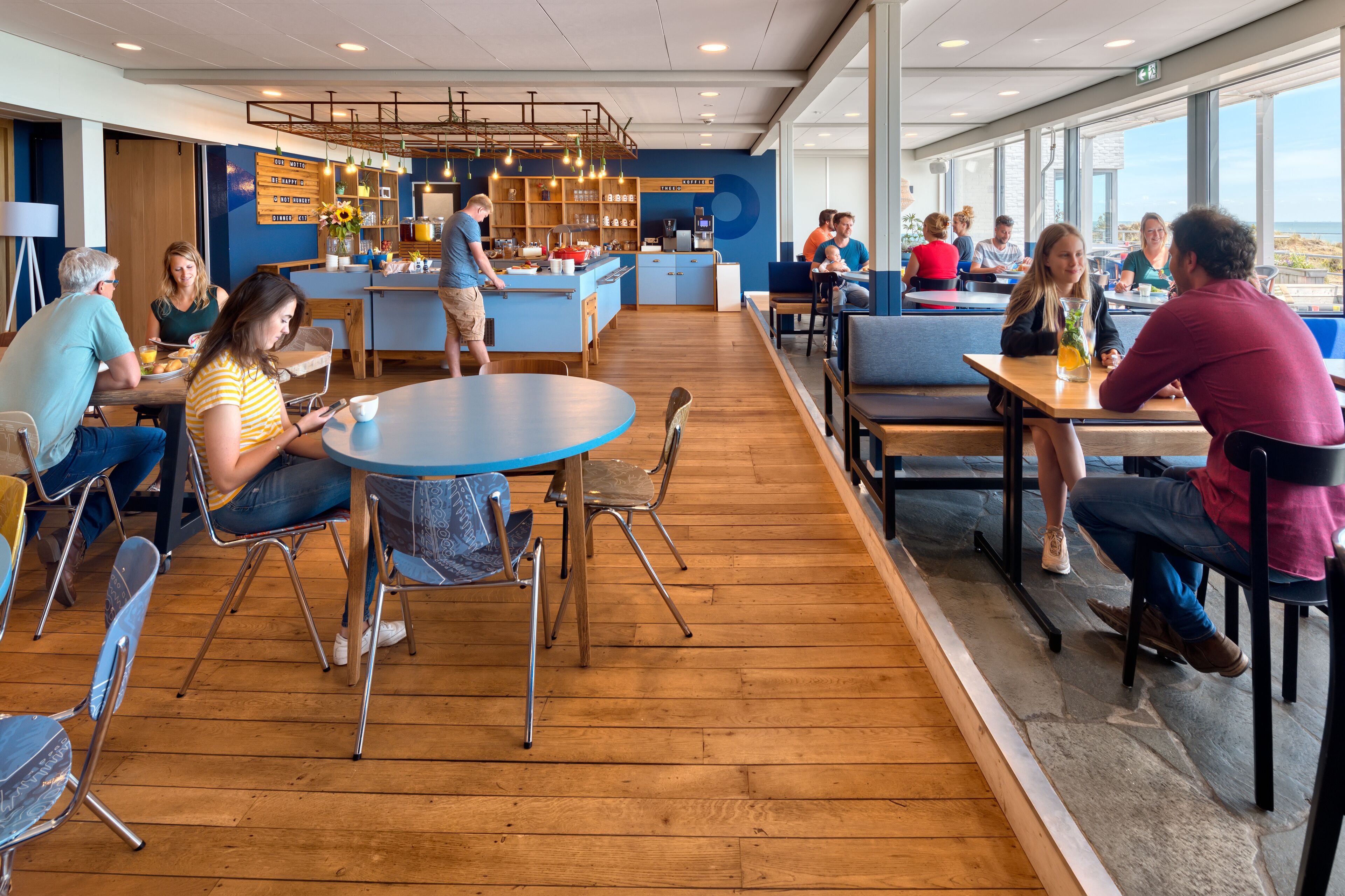 Een moderne eetruimte met houten vloeren, ronde tafels en metalen stoelen. Er is een keuken met een aanrecht en diverse keukenbenodigdheden.