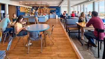 Een moderne eetruimte met houten vloeren, ronde tafels en metalen stoelen. Er is een keuken met een aanrecht en diverse keukenbenodigdheden.