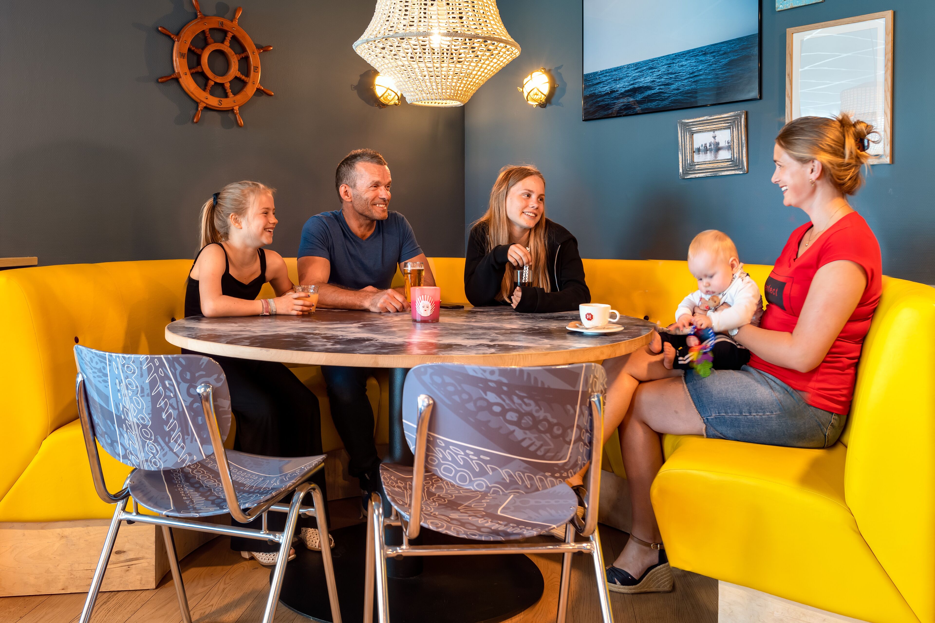 A family dining at a table with a round table and chairs, in a room with a yellow couch and a ship's wheel decoration on the wall.