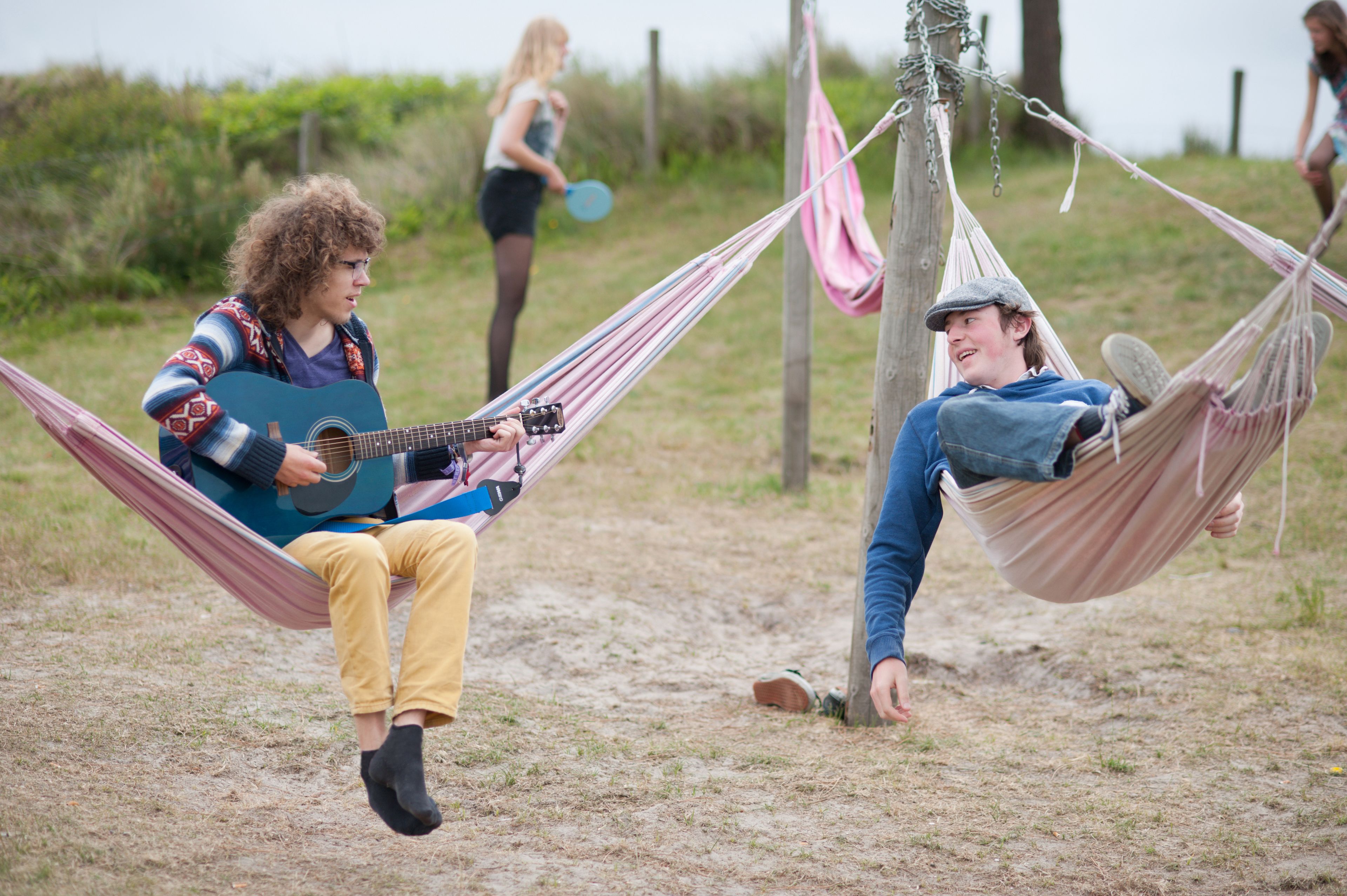 Two people relaxing in hammocks on a sandy beach with a guitar player and another person in the background.