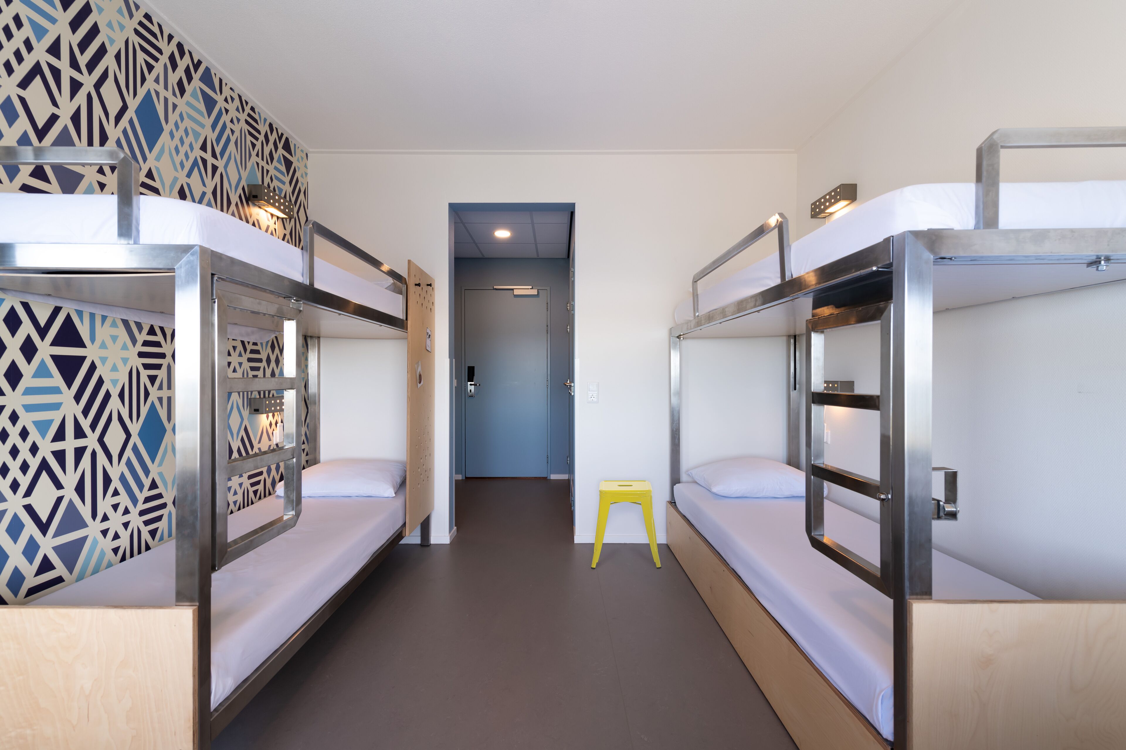 A corridor with bunk beds, a door, and a small yellow stool.