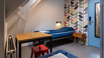 A small, well-lit room with a bed, a table, and a red stool. There is a radiator on the left and a door with a colorful pattern on the right.
