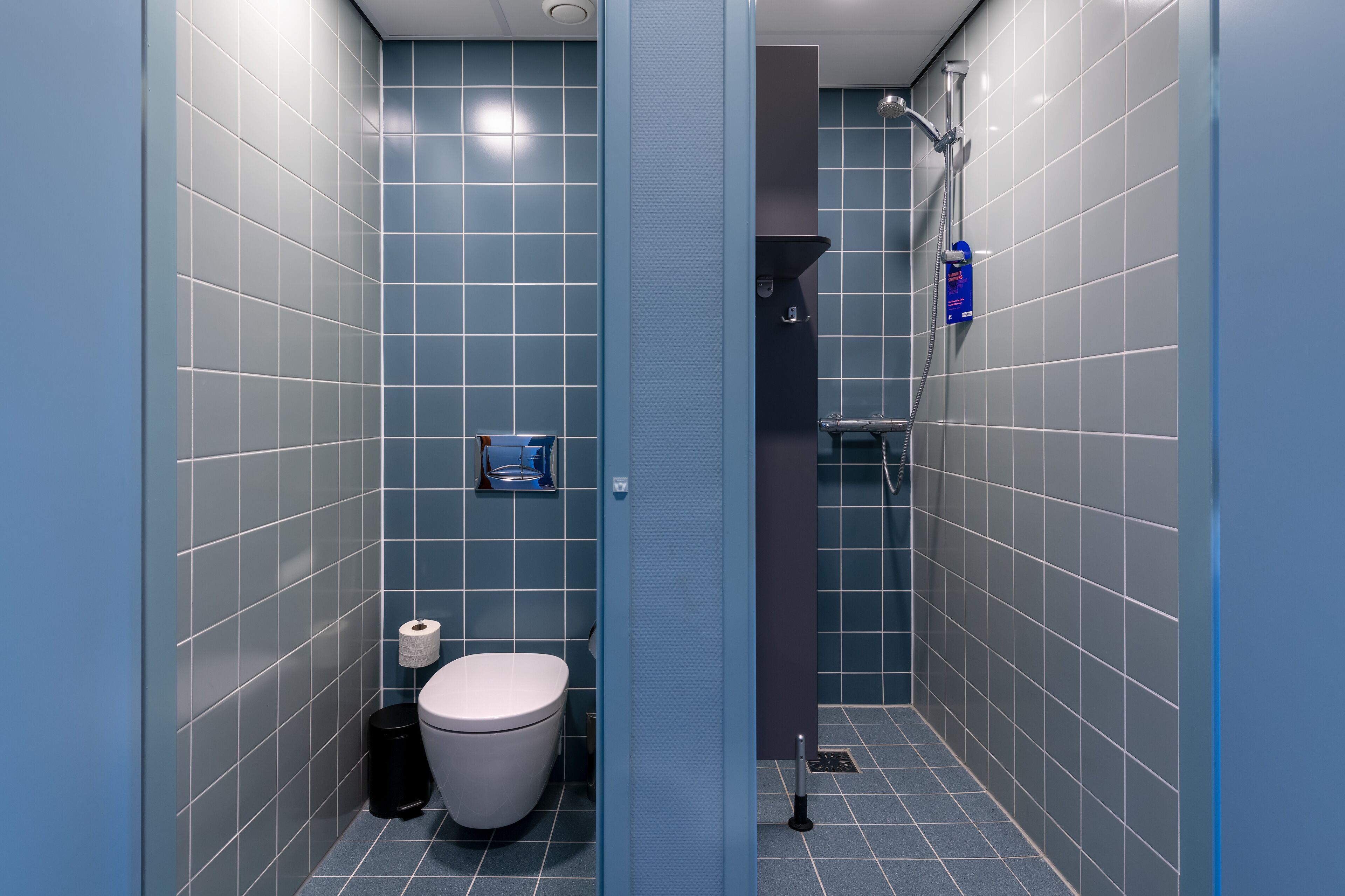 A modern bathroom with blue tiles, a toilet, a trash bin, and a shower area.