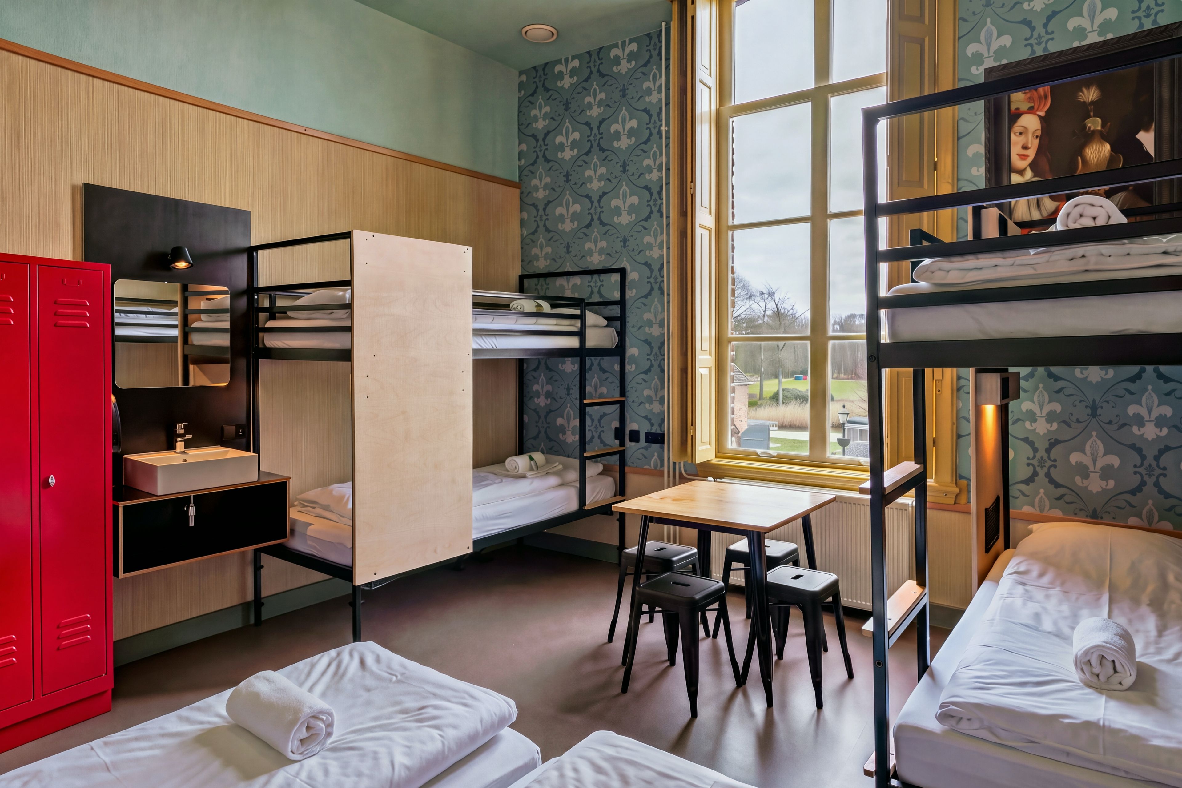 A dormitory room with bunk beds, a desk, a chair, a red wardrobe, and a window with a view.