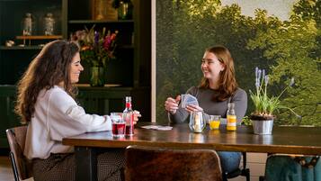 Two people playing cards at a wooden table with drinks and a potted plant.