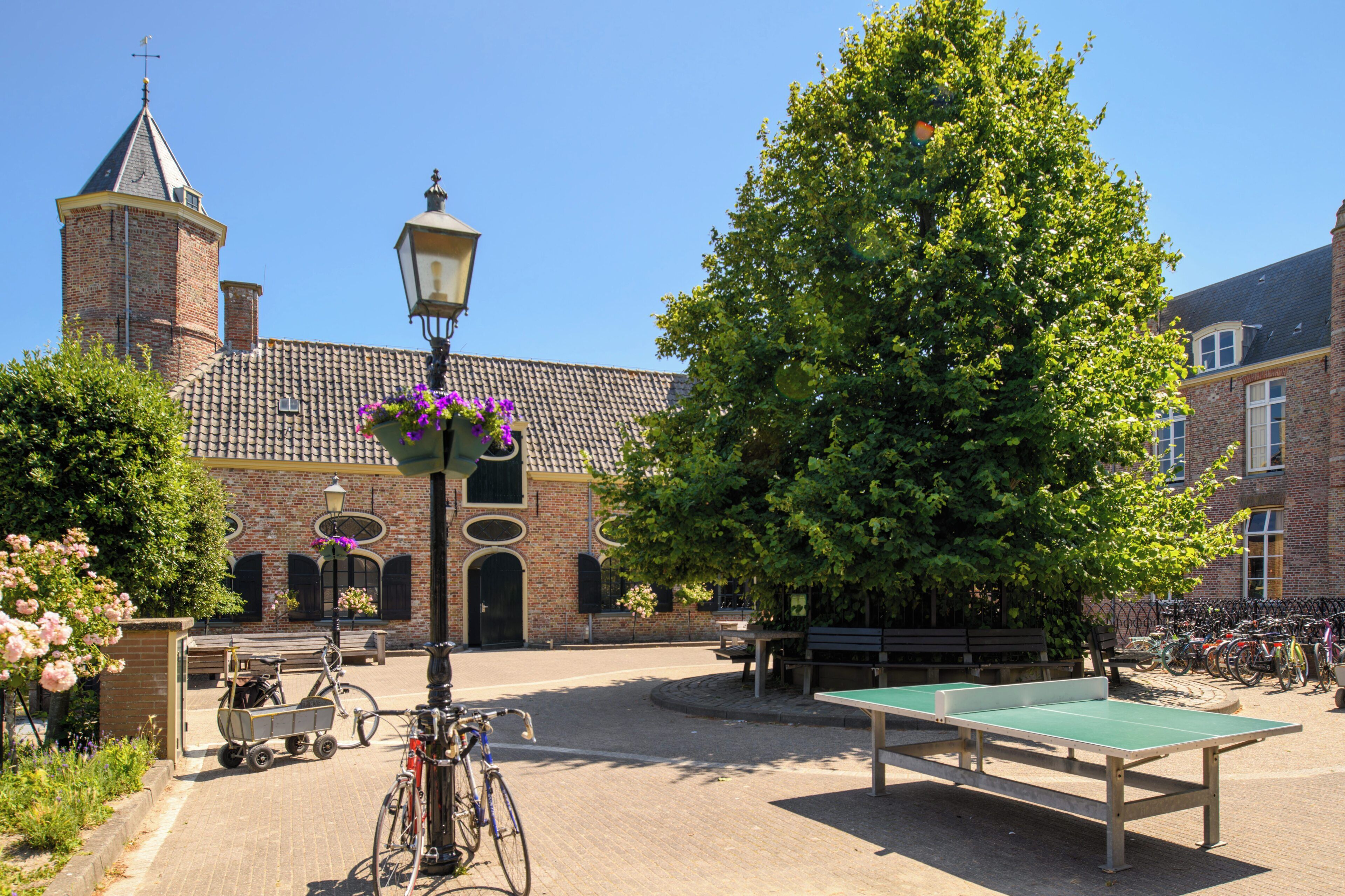 A courtyard with a ping pong table, bicycles, a bench, and a building with a brick facade and a tower.