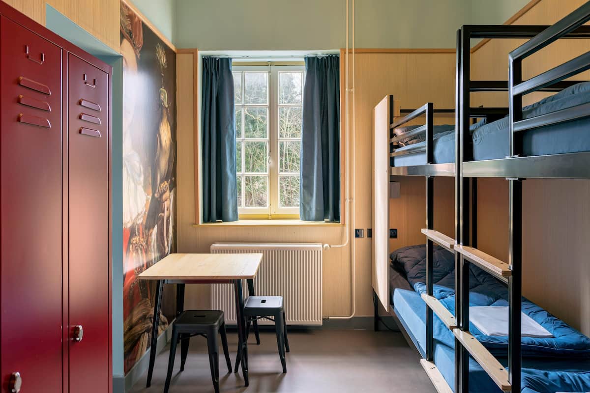 Een studentenkamer met stapelbedden, een bureau, een stoel, een radiator en een raam met gordijnen.