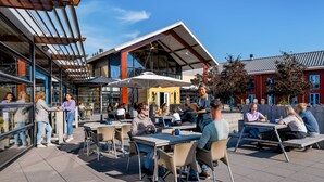 Un café en plein air avec des tables et des chaises, un bâtiment moderne doté de grandes fenêtres, et des gens qui déjeunent et discutent.