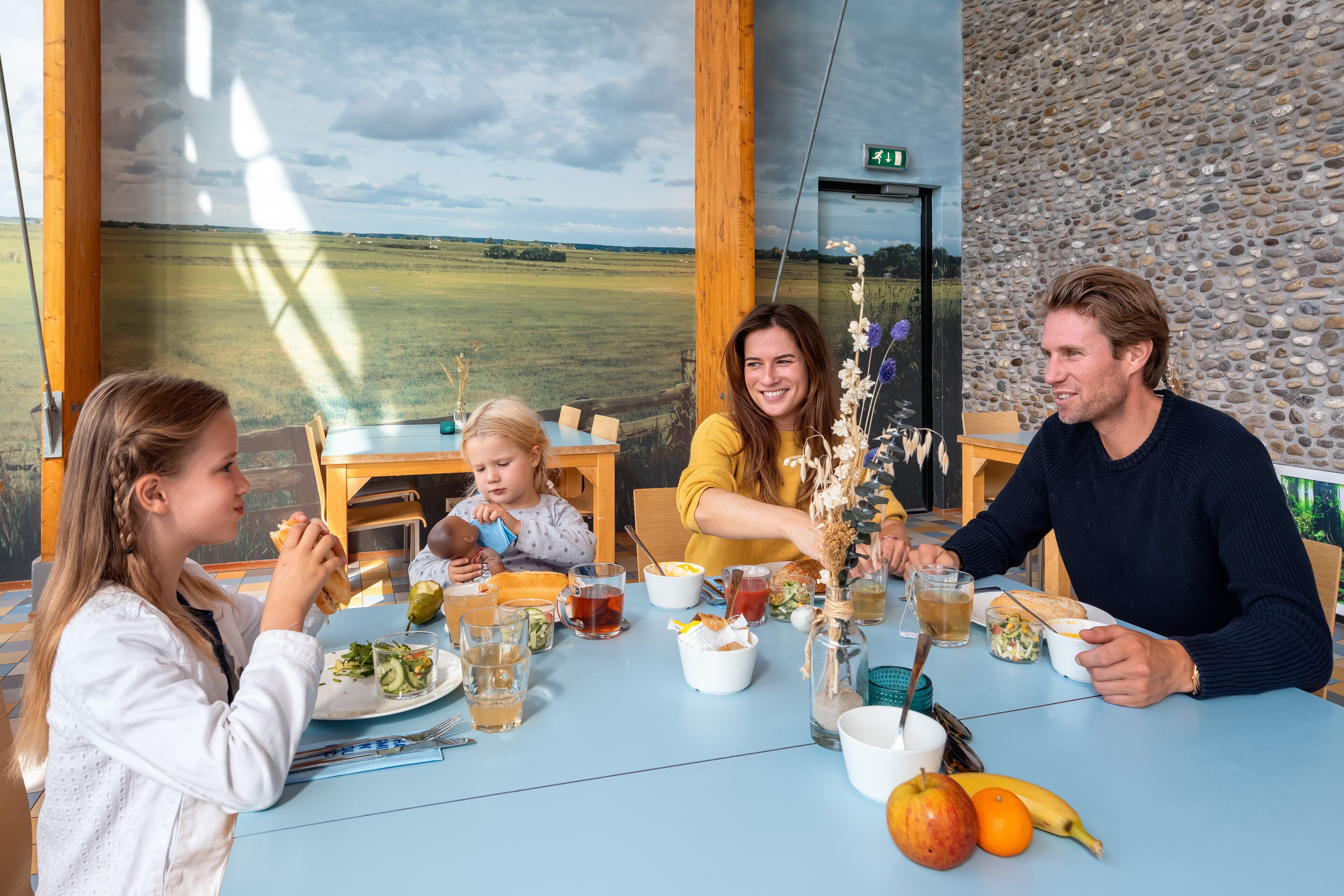 A family dining at a table with a view of a field, a stone wall, and a wooden ceiling.