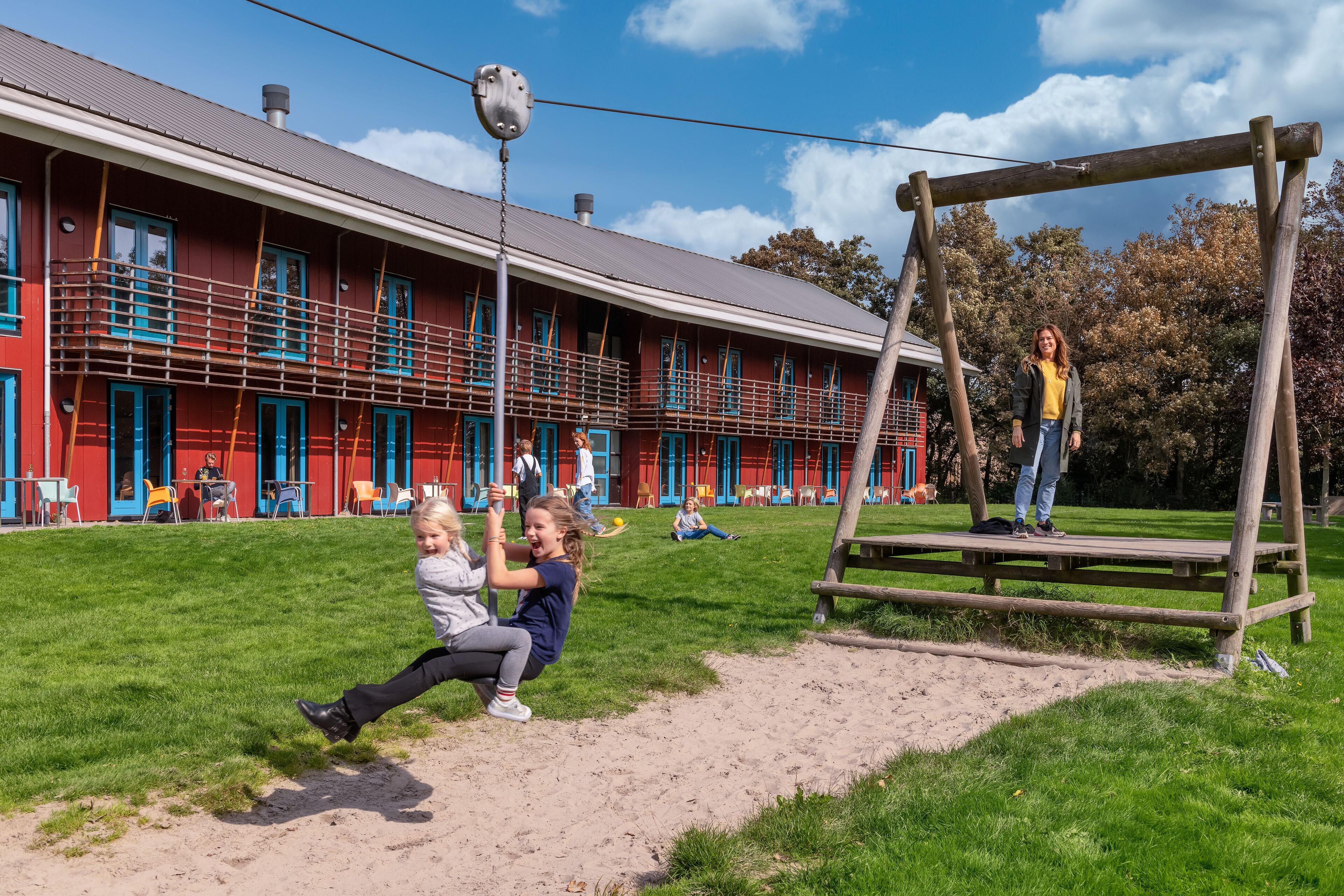 Un parque infantil con columpios y un tobogán, un edificio rojo con puertas y ventanas azules, y un área verde con gente.