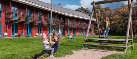 Un parque infantil con columpios y un tobogán, un edificio rojo con puertas y ventanas azules, y un área verde con gente.