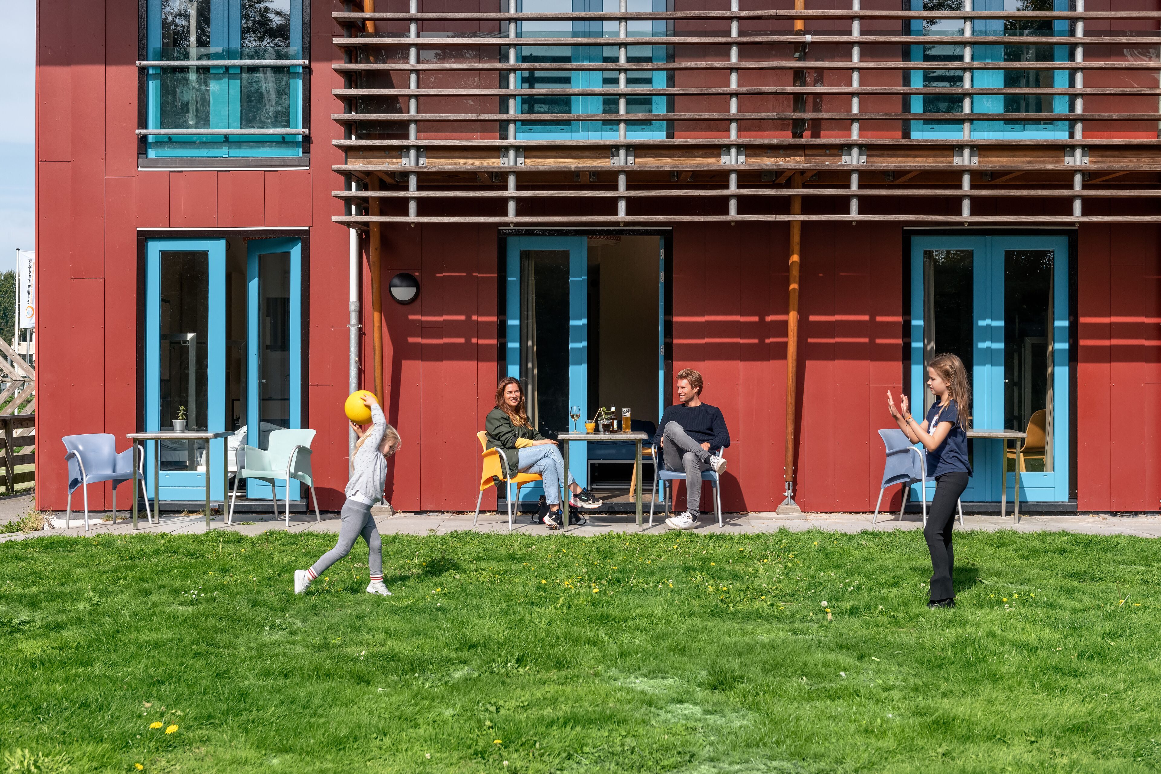 A modern building with red exterior walls and blue accents, featuring outdoor seating and a grassy area.