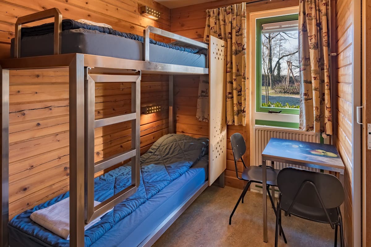 Habitación con literas, un escritorio, una silla, paredes de madera, una ventana con cortinas y un radiador.