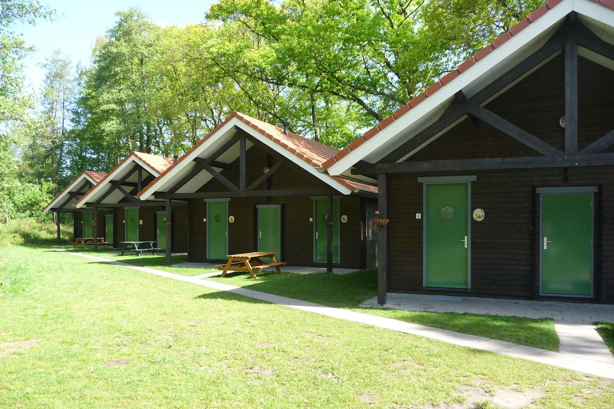 Una hilera de cabañas de madera con puertas verdes, cada una con una mesa de picnic y un banco.