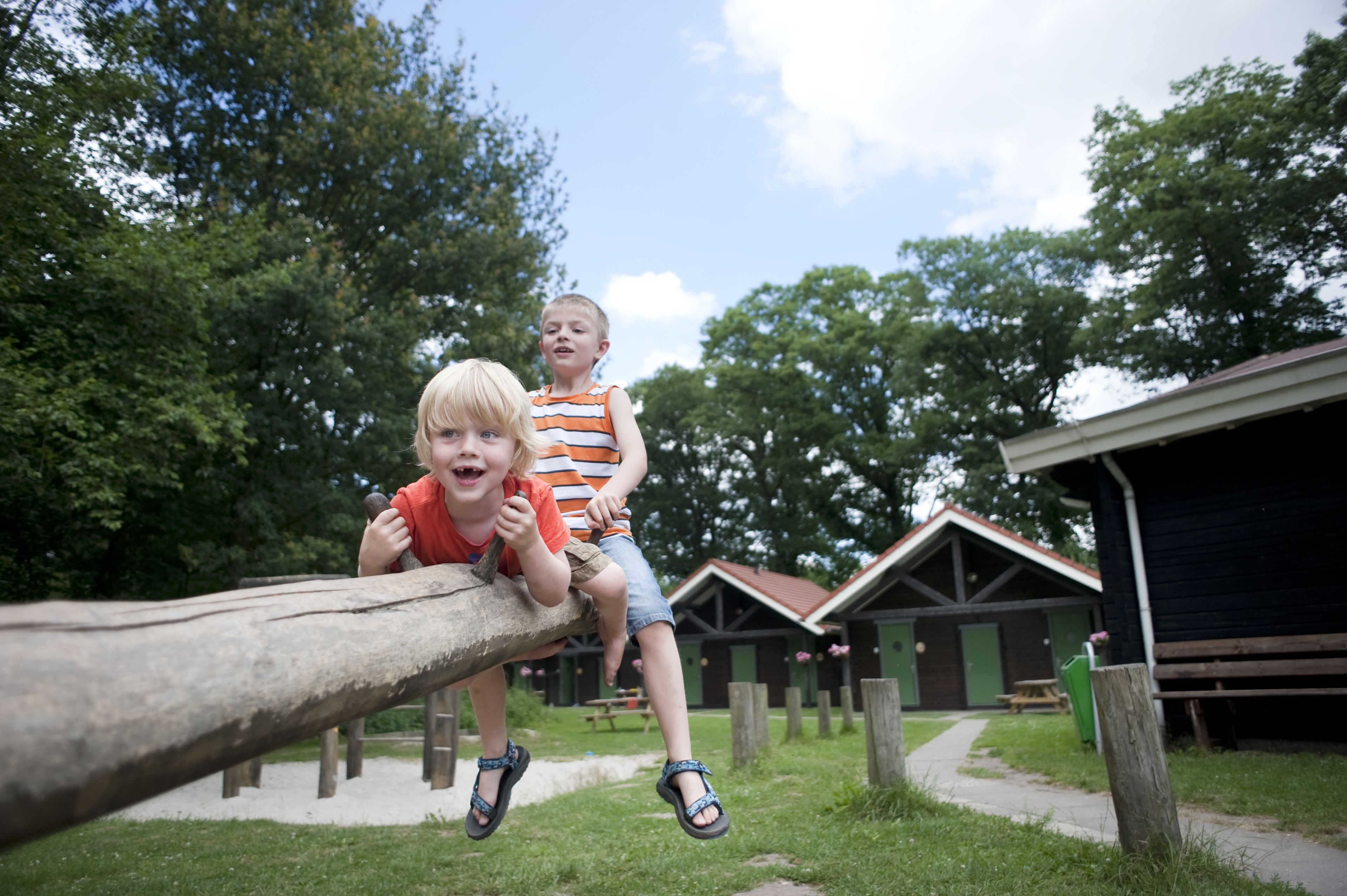 Dos niños jugando en un puente de tronco en un parque con mesas de picnic y cabañas al fondo.