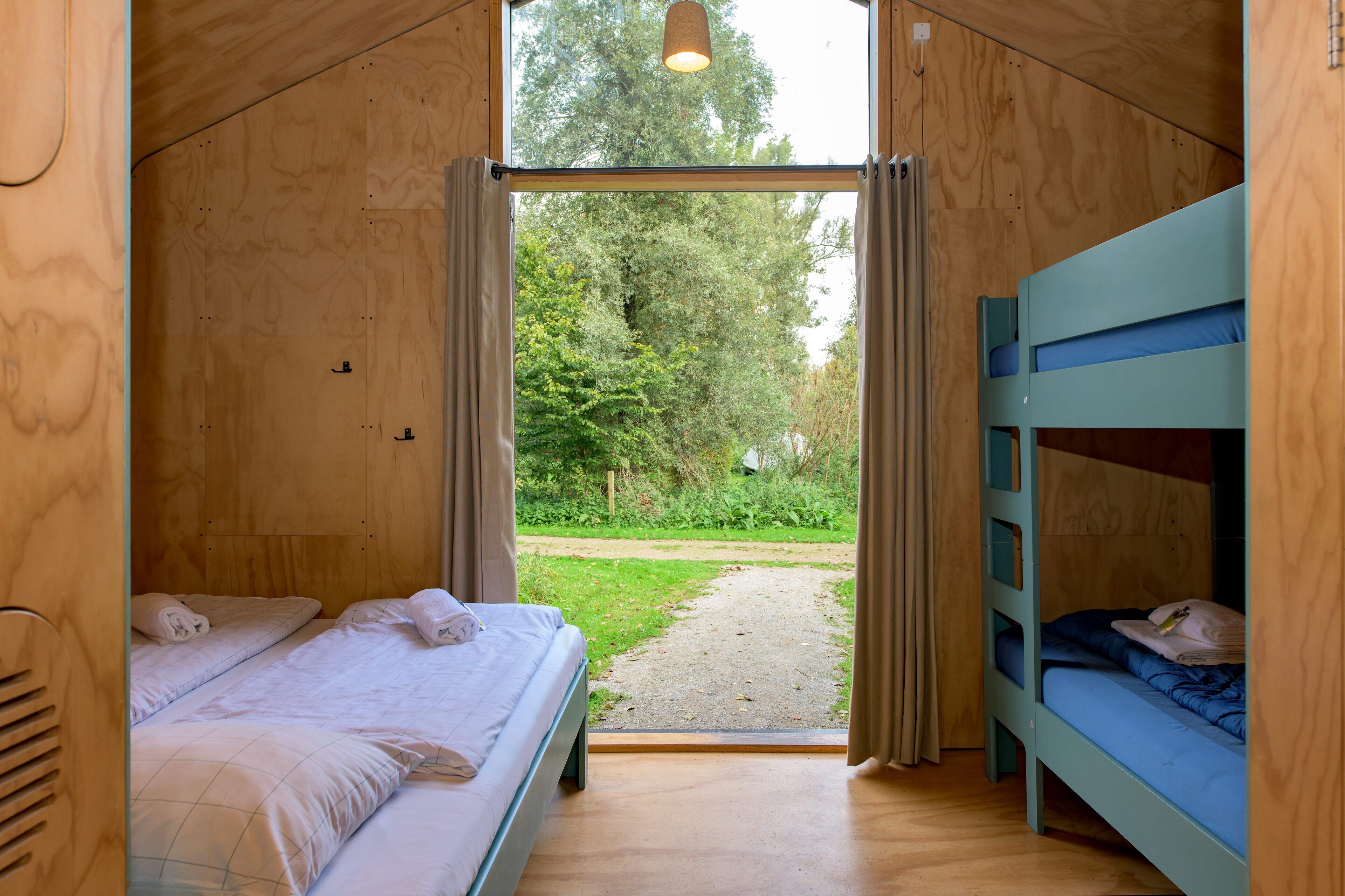 Ein Holzinnenraum mit einem Bett, einem Etagenbett und Blick auf eine grüne Landschaft durch eine offene Tür.